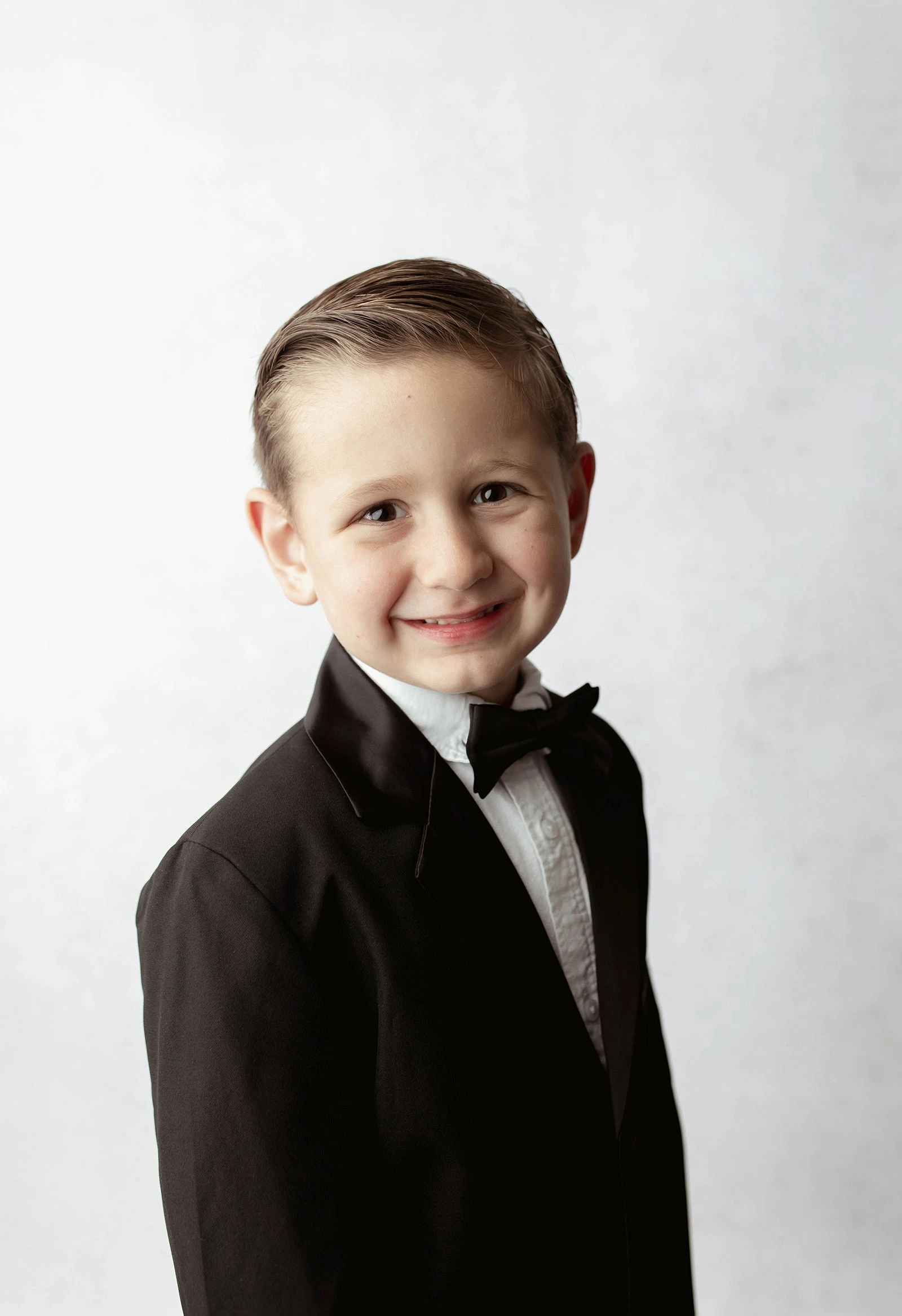The image shows a formal studio portrait of a young child framed from the chest up against a light, smoothly textured background. The child wears a black suit jacket, white dress shirt, and black bow tie. Hair is neatly combed, and lighting is soft and even, creating a polished, traditional portrait style. The image shows a formal studio portrait of a young child framed from the chest up against a light, smoothly textured background. The child wears a black suit jacket, white dress shirt, and black bow tie. Hair is neatly combed, and lighting is soft and even, creating a polished, traditional portrait style.