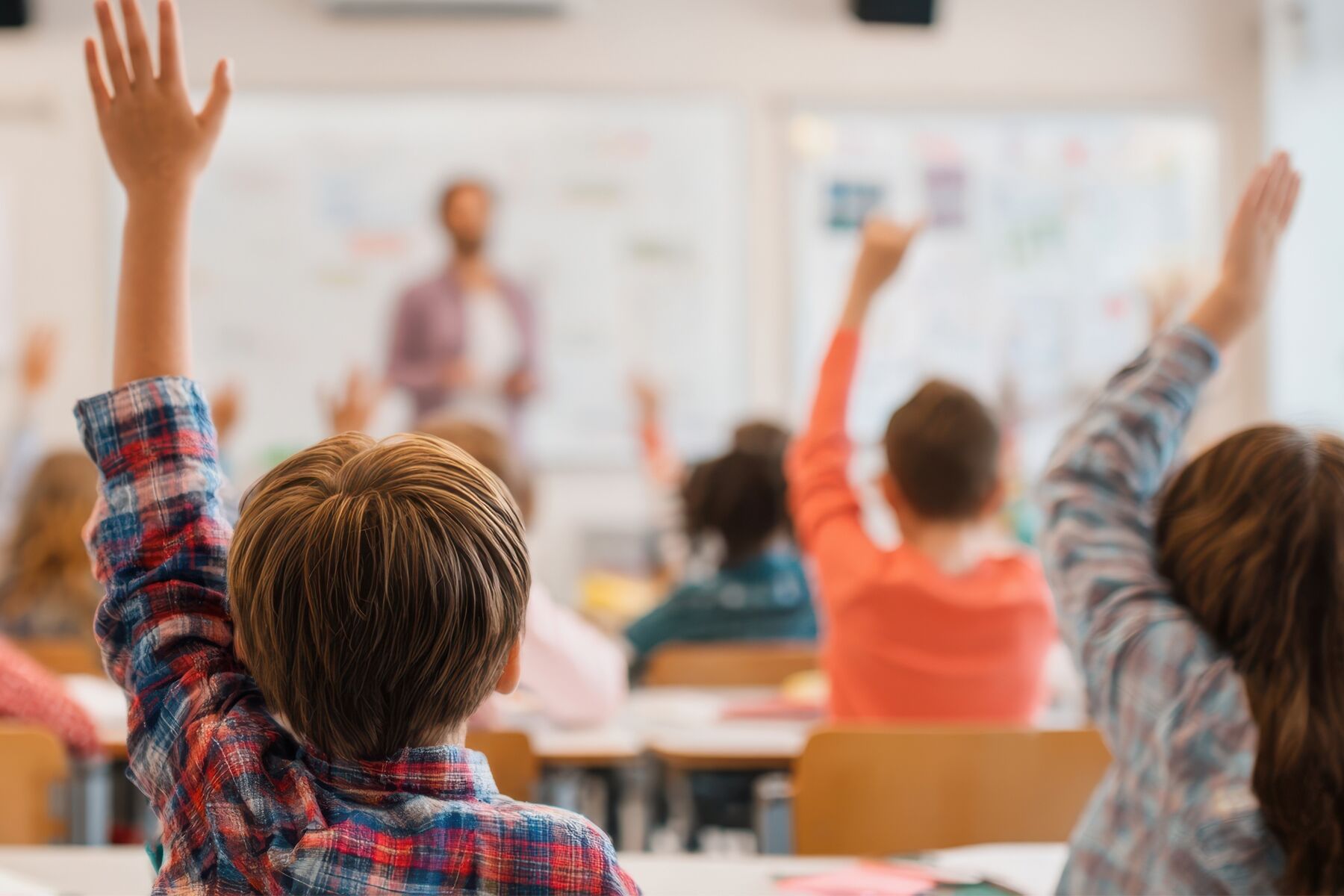 Students seated in a classroom raise their hands while a teacher stands at the front near a whiteboard