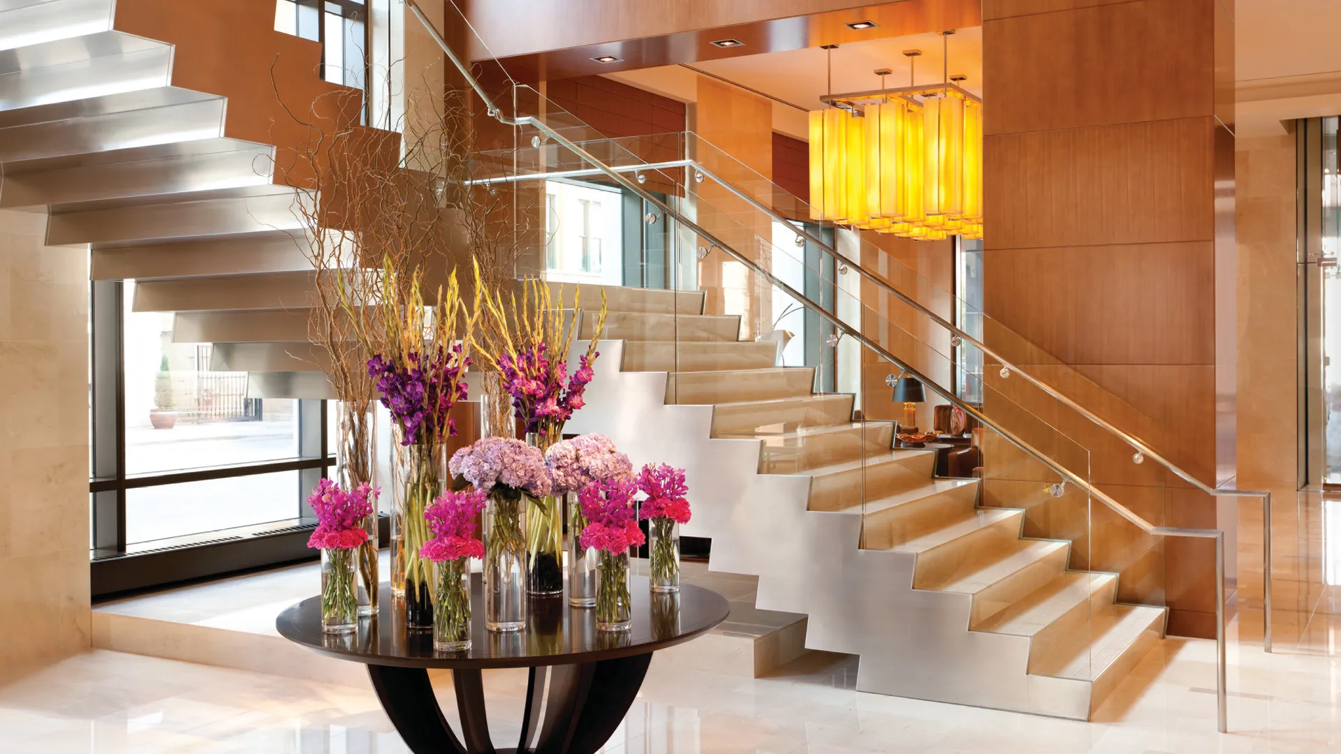 Modern interior with a floating staircase, glass railing, warm wood walls, hanging amber lights, and a round table holding tall colorful floral arrangements Modern interior with a floating staircase, glass railing, warm wood walls, hanging amber lights, and a round table holding tall colorful floral arrangements